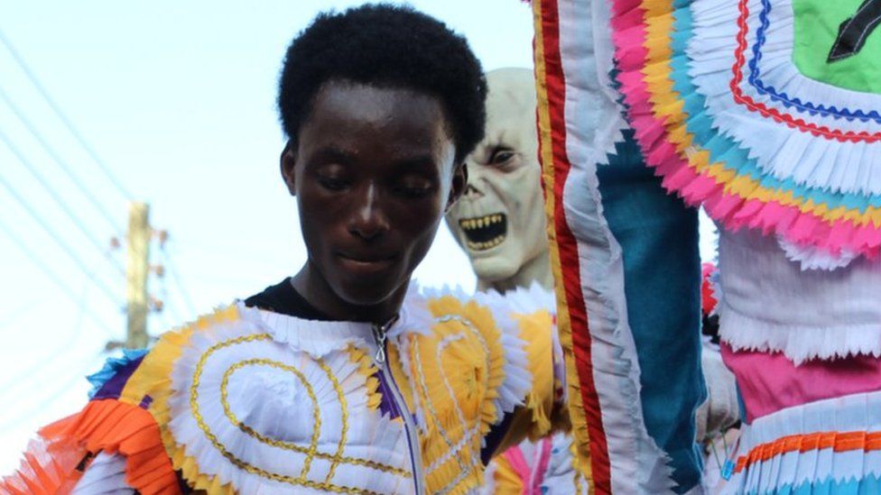 Man dressed in costume made with multi-coloured, chequered fabric. Lurking behind him, someone in an alien costume in Sekondi Ghana
