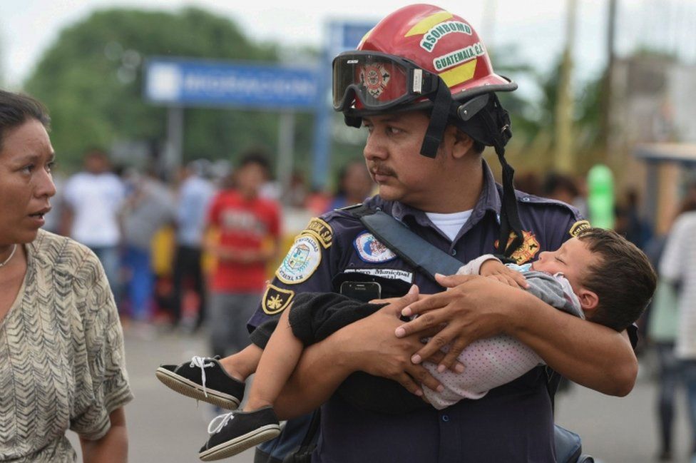 A Guatemalan firefighter carries an ailing toddler while speaking to a woman, possibly the boy's mother