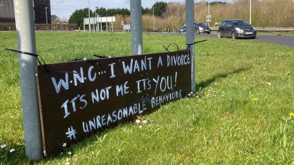 Black sign with white lettering on a roundabout. The sign says: "WNC... I want a divorce"