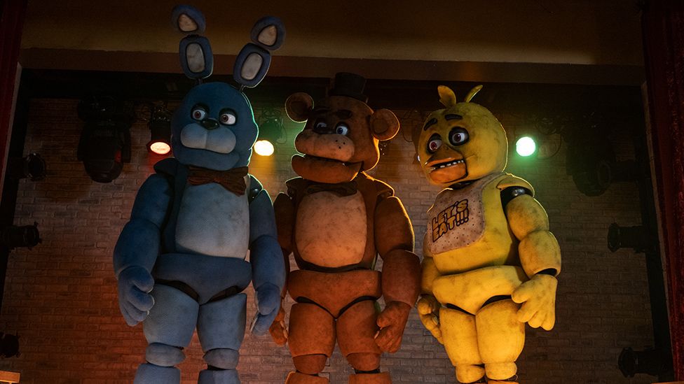 Three animatronic creatures stand on a stage with spotlights behind them. They are cartoonish in appearance. On the left is a light blue rabbit with a red bow tie. In the middle is a teddy bear wearing a top hat. And on the right is a bird-like character, who's yellow, wearing a bib with "Let's Eat!" written in capital letters. Their expressions are neutral but there's an air of menace about them.