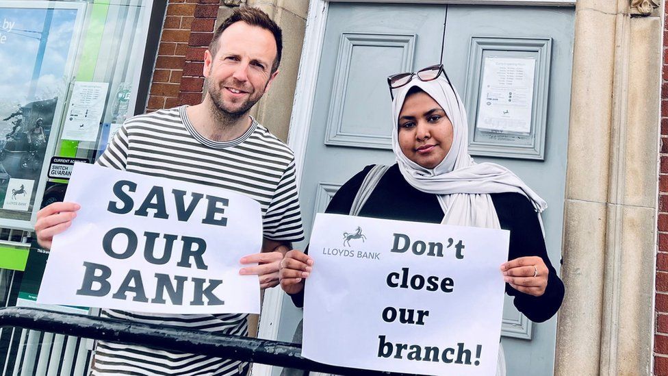Councillors Ben Miskell and Nabeela Mowlana standing outside Lloyds Bank holding placards