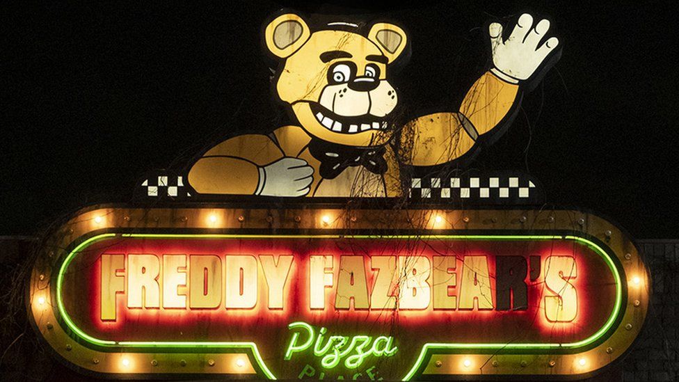 A slightly grubby neon sign that reads "Freddy Fazbear's Pizza" in block capital letters. Above the lettering is an illuminated bear character. He's smiling and has one arm raised in a wave.