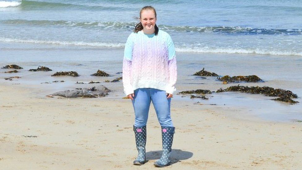 Emma is standing on a beach with the sea behind her wearing a rainbow jumper jeans and wellington boots