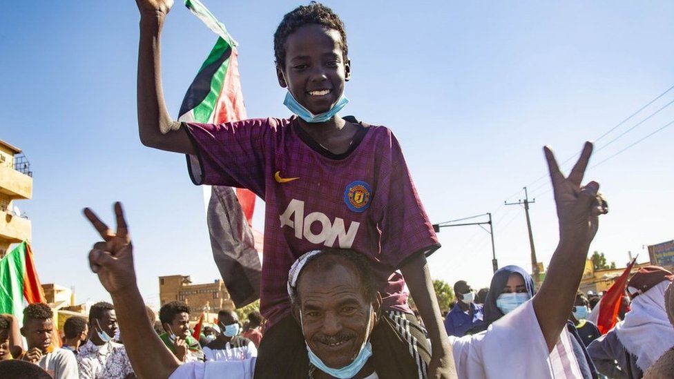A man smiling making the victory sign. He is carrying a boy on his shoulders who is holding the Sudan flag.