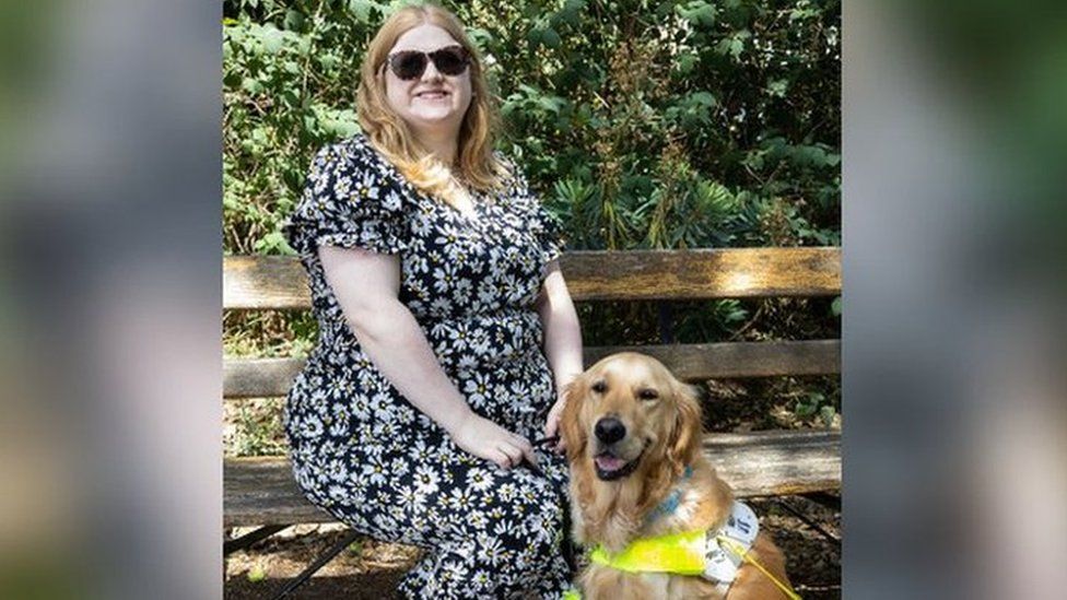 Dr Amy Kavanagh sitting on a bench wearing a black and white dress, with her guide dog Ava who is wearing a yellow harness.