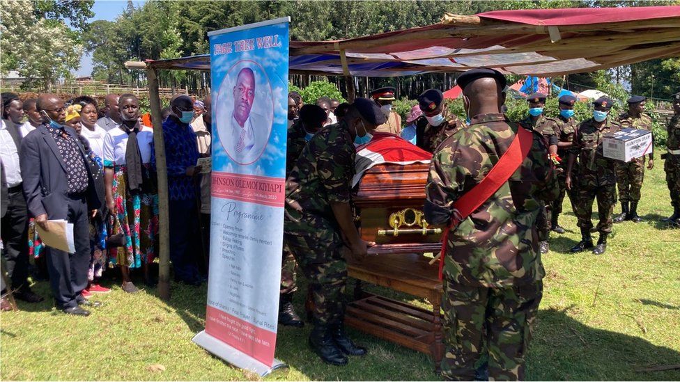 Funeral service, with a photo of the dead soldier and attendees as well as soldiers standing around the casket.
