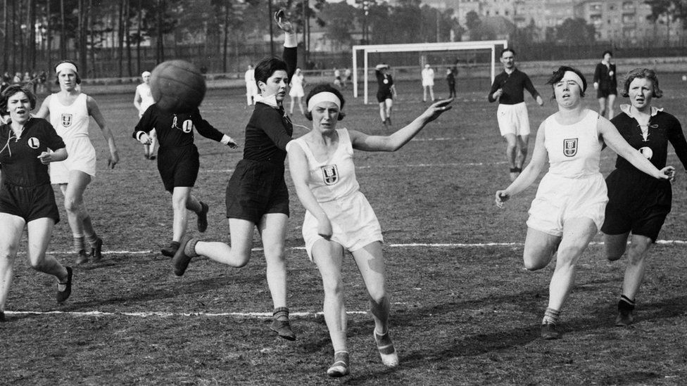 Two groups of women wearing black and white mid-length sportswear enjoy a handball game in a Berlin park 1935.