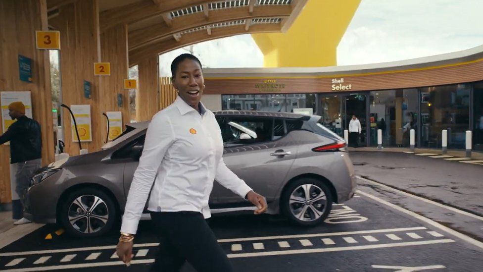 A member of Shell staff walking across a Shell forecourt with electric vehicles charging in background