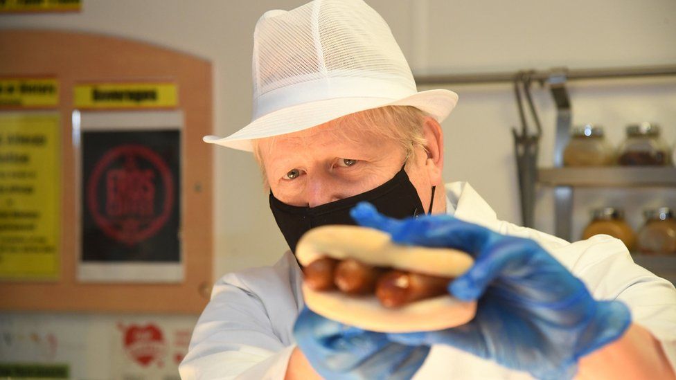Prime Minister Boris Johnson holds up a sausage filled roll as he assists kitchen staff during a visit to Royal Berkshire Hospital, Reading, to mark the publication of a new review into hospital food