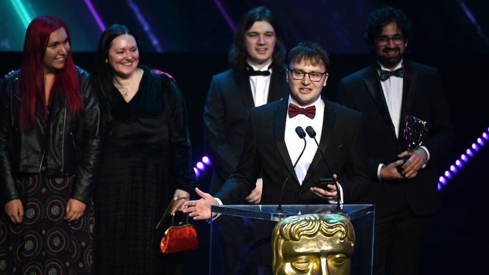 A man in glasses, black tuxedo with a white shirt and red bow tie stands with his hands at his sides, palms up, as he speaks into a microphone on the Bafta lectern. The lectern bears the golden mask of the Bafta Academy - a male face with carved features. Behind him, a woman with red hair, woman with black hair, man with shoulder-length black hair in a tuxedo and a man holding the award at waist height stand in a row behind him. They're all smiling, joining in the joy of winning an award.