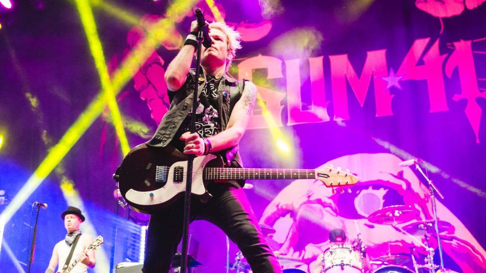 Deryck Whibley of Sum 41 performs on stage during day 3 of Download festival 2019 at La Caja Magica on June 30, 2019 in Madrid, Spain. Deryck is a white man in his 40s with bleached blonde hair. He's wearing a sleeveless black denim jacket over a black top, paired with tight black jeans. He has a guitar slung over his shoulder and holds a microphone with his right hand. The staging is lit by pink and yellow lights with the band's logo behind Deryck.