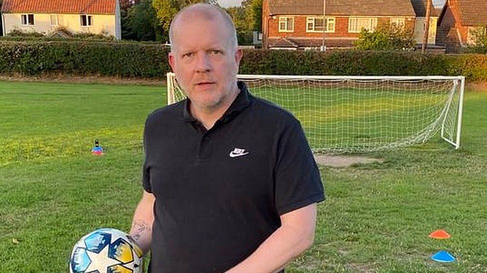 Simon Compton holding a football wearing a black T-shirt and black shorts