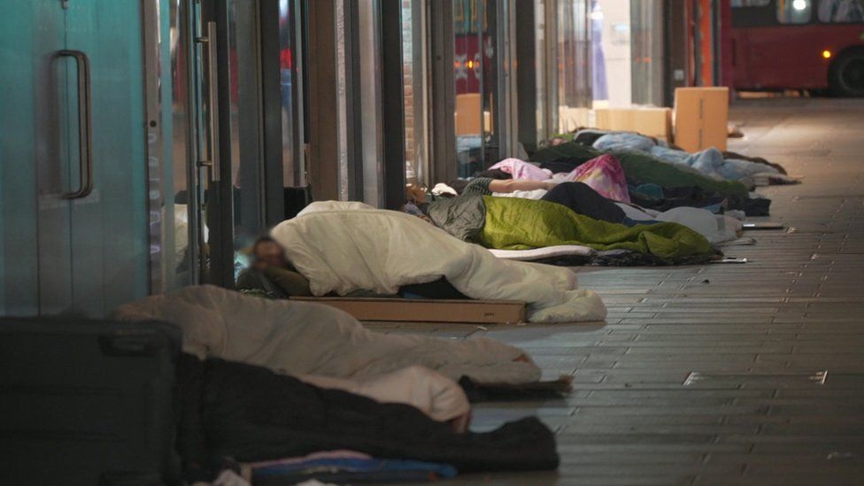 Rough sleepers in sleeping bags lie on the street outside a row of buildings
