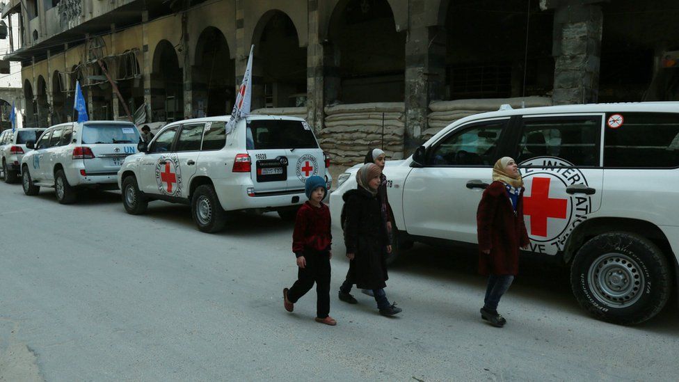 Syrian children walk past vehicles of the UN and the International Committee of the Red Cross (ICRC) delivering humanitarian aid in the Syrian town of Douma