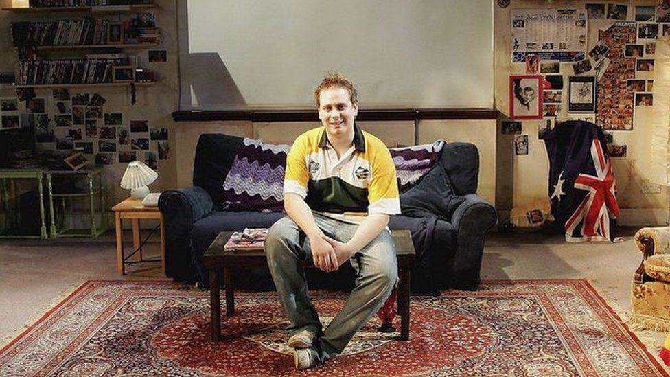 Patrick Harvey sitting on a sofa in living room of Neighbours set