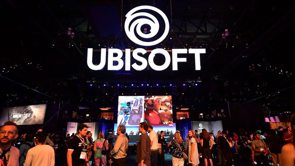 Огромная вывеска Ubisoft висит в воздухе на оживленном торговом зале, заполненном людьми и демонстрирующим игры