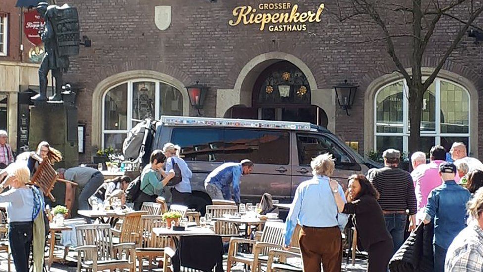 Picture of van in square with statue and crowd surrounding it, outside of restaurant.