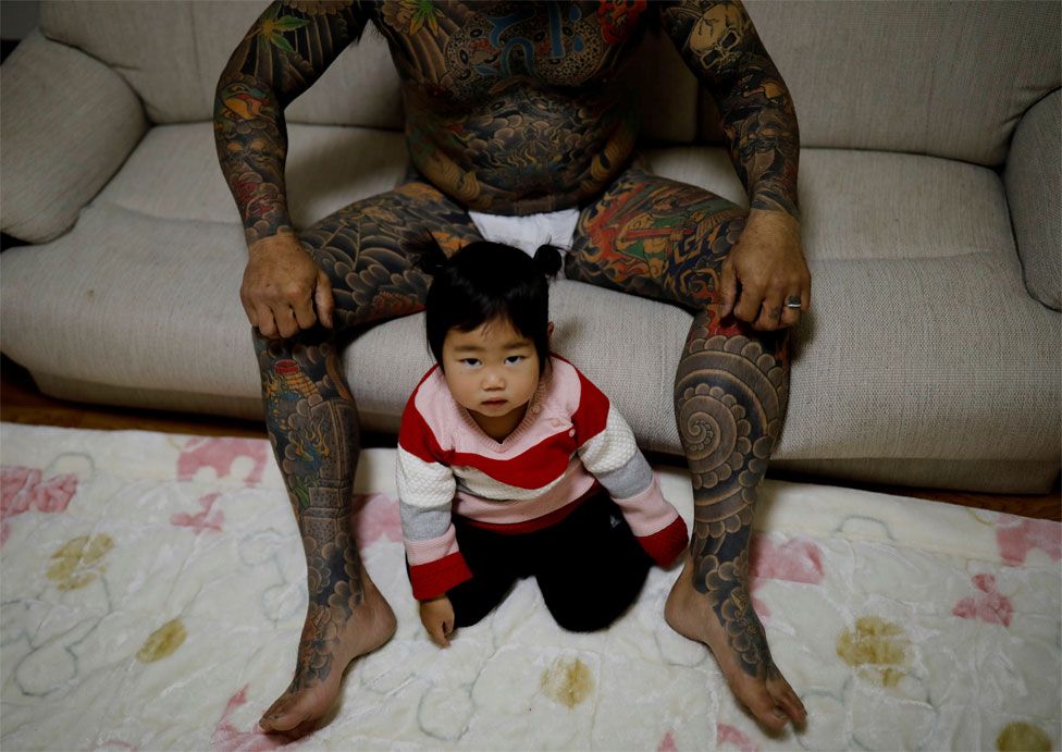 A man with tattoos on his legs and torso poses with this baby daughter sat on the carpet