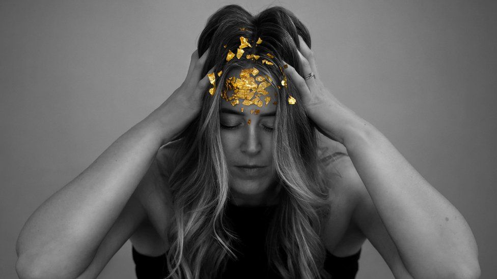 Black and white image of a woman with her hands on the side of her head. The only form of colour in the picture is the gold sequins on her forehead.