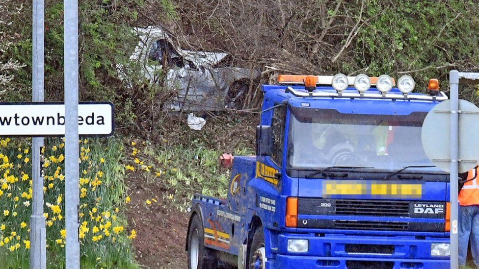 A silver Mercedes car crashed in undergrowth, with a vehicle recovery lorry parked nearby