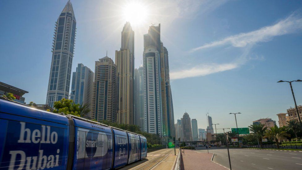 A tram with the words 'hello Dubai' passes in front of the Dubai skyline.