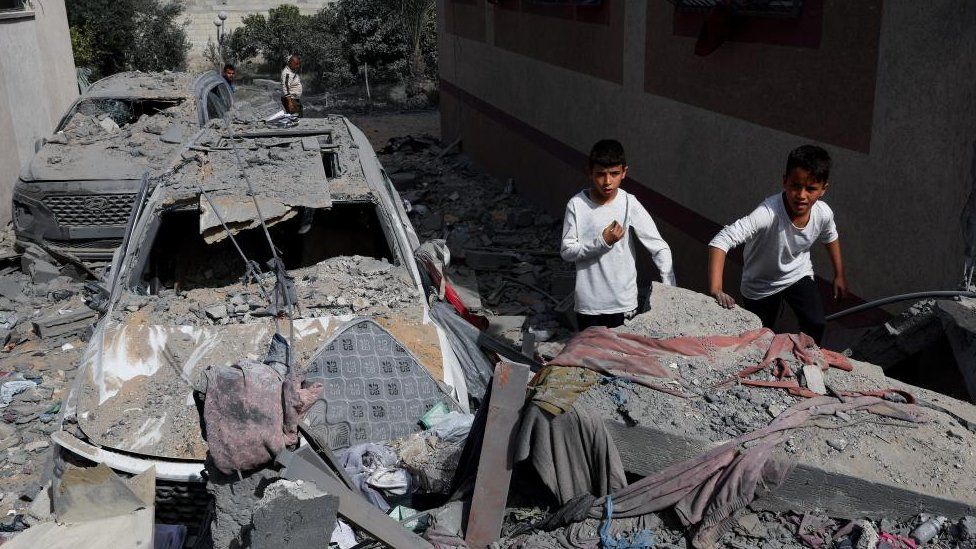 Two boys seen stood next to a car that is heavily damaged and covered in debris in Gaza. In the foreground is what appears to be the rubble of a collapsed building.