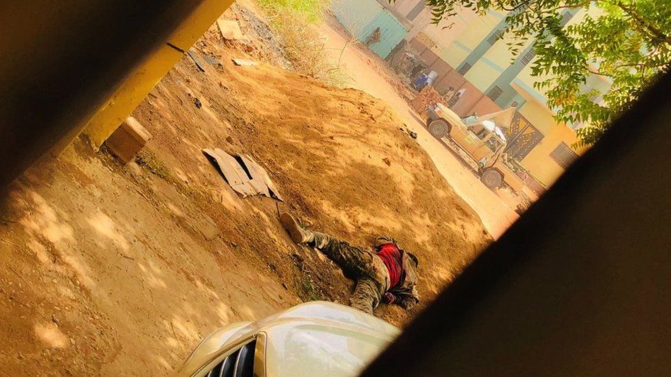 Тело солдата лежит рядом с насыпью земли, в то время как солдат использует здание в качестве укрытия через улицу в Хартуме 15 апреля 2023 года, поскольку боевые действия обострились