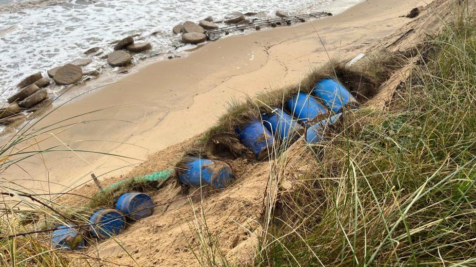 Plastic barrels and marram grass on beach cliff.
