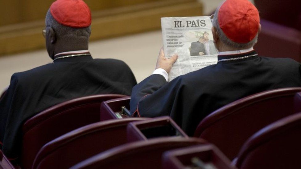Spanish Cardinal Ricardo Blazquez Perez (right) reads a newspaper showing a picture of gay bishop Krzysztof Charamsa and his partner Eduard before the start of the morning session of the Synod of bishops on family issues at the Vatican (09 October 2015)