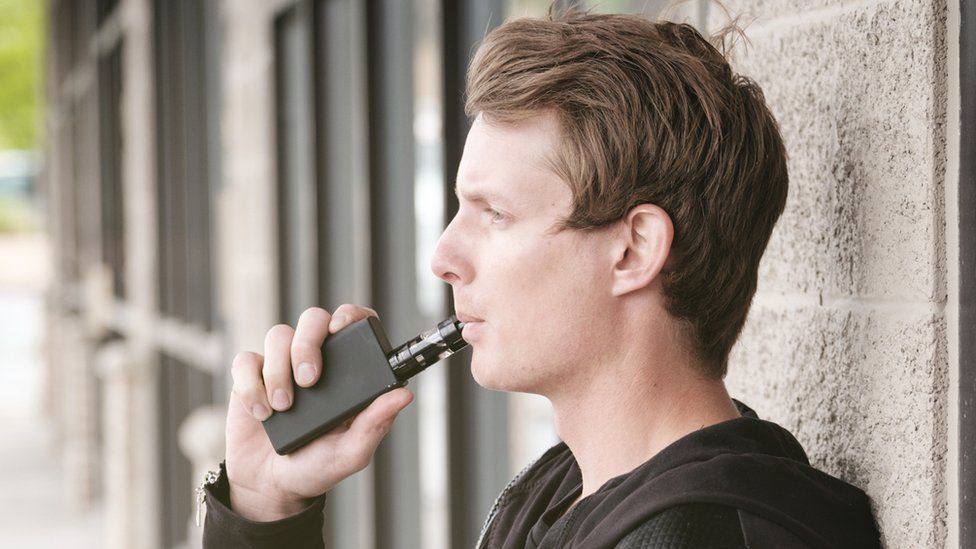 A man leans against a grey concrete column outside an office or college building. He's wearing a black hoodie. In one hand, he's holding an electronic cigarette and pressing the device to his lips. It's black, with a square body and cylindrical e-liquid tank.