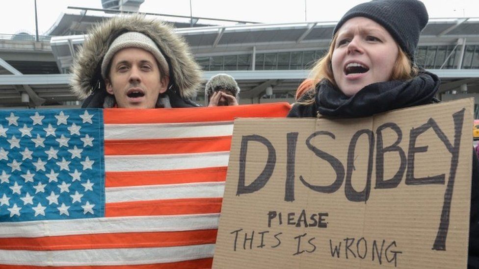Demonstrators with a US flag and a sign urging protest stand at JFK airport