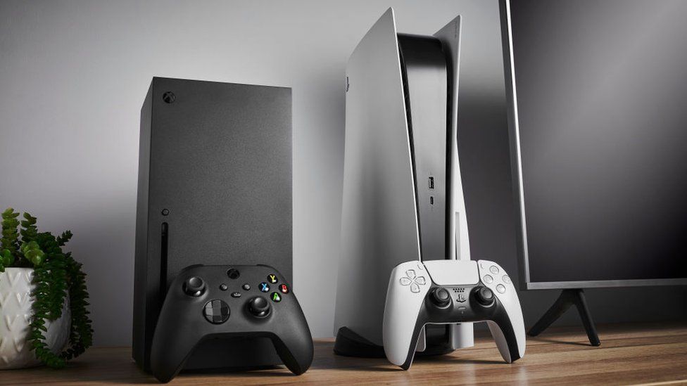 An Xbox Series X and Playstation 5 side-by-side on an office desk that's pristine and clean. Both machines are standing vertically, with their respective controllers propped against them. A computer monitor is partially visible on one side of the image and a small potted plant on the other.