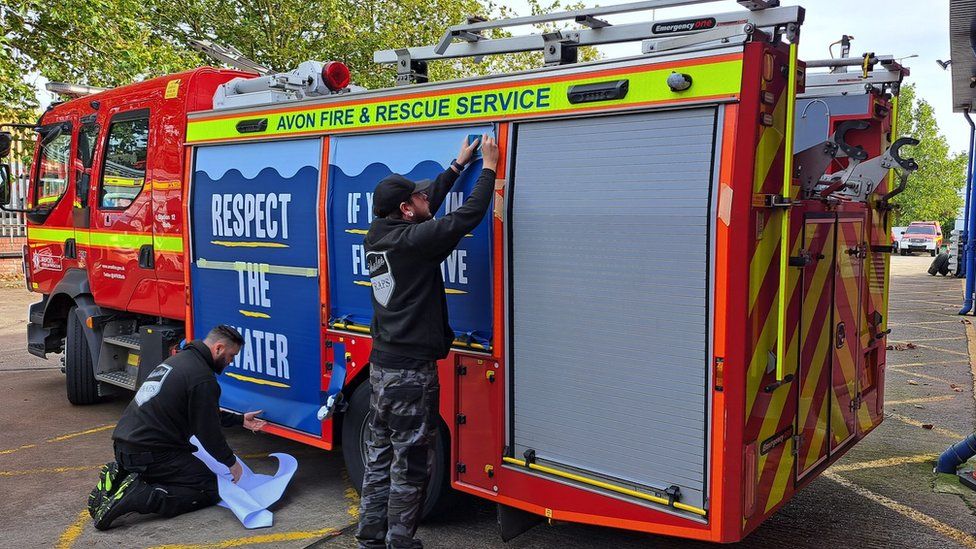 Two men putting the new wrap onto a fire engine. Their backs are to the camera. Both panels are royal blue with white capital letters which are underlined in yellow. The first panel reads 'Respect the water' and the second reads 'If you fall in, float to live'.