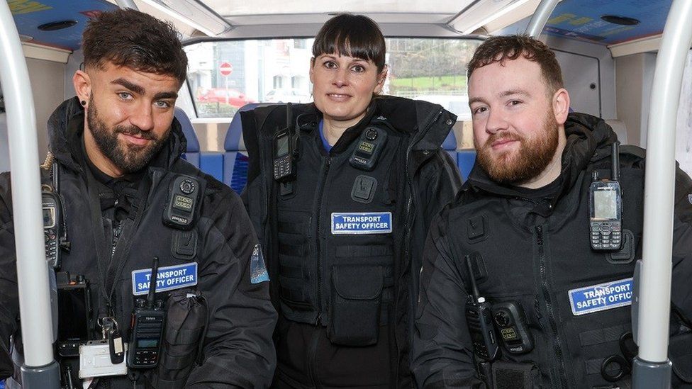 Three Transport Safety Officers on a bus - two men and a woman wearing black uniform coats with personal radios attached to the lapel area.