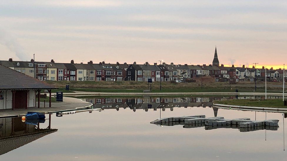 A boating pond with houses beyond reflected on the water