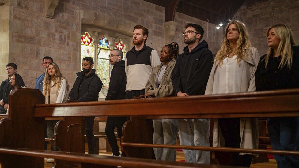 The cast of the Traitors lined up in a church. L-R Harry, Charlotte, Andrew, Jaz, Zack, Paul, Jasmine, Ross, Charlie and Evie. The cast stand in pews inside a stone church with stained glass windows visible behind them
