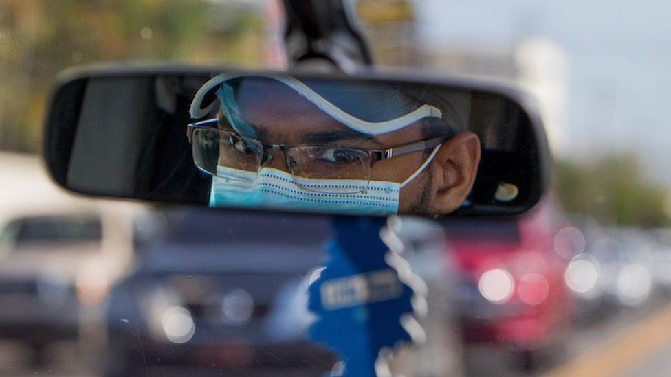 A Uber driver wears a face mask as seen in his rearview mirror