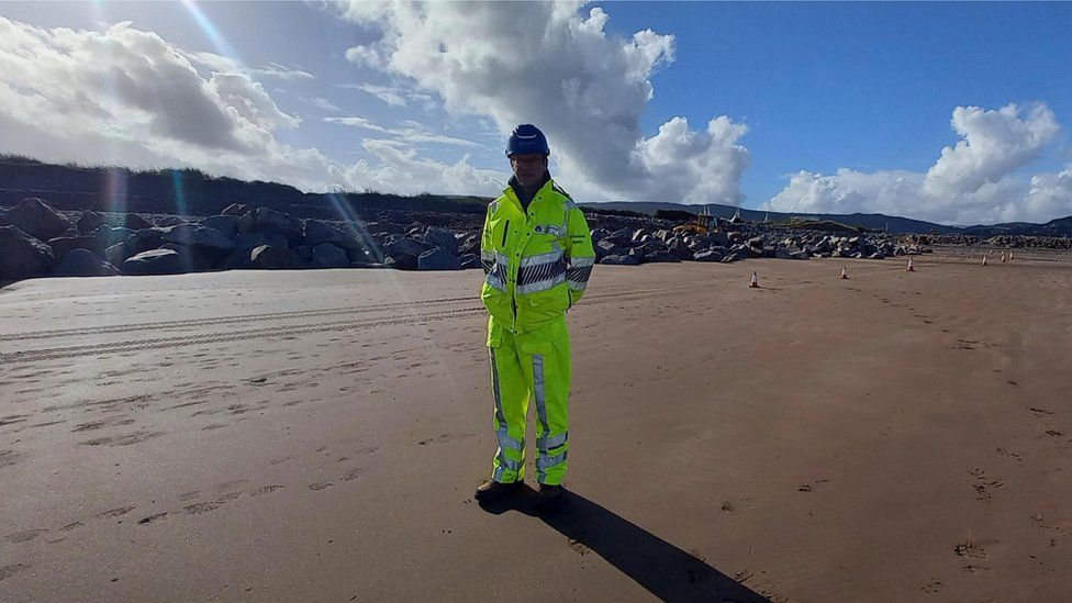A man in a hard hat and full high vis outfit standing on a sunny sandy beach