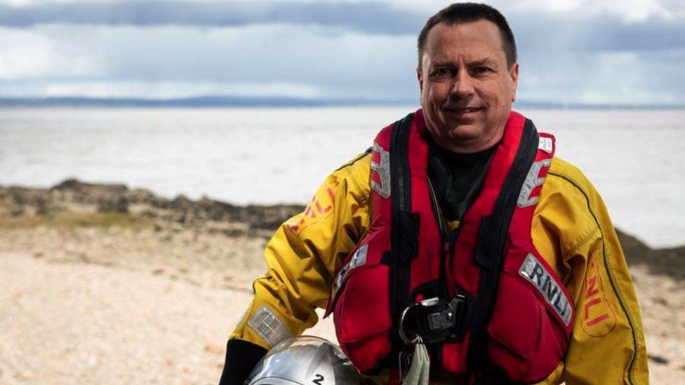 Bruce du Preez. He is stood near the coast, wearing his RNLI uniform. It is yellow and includes a red life jacket.