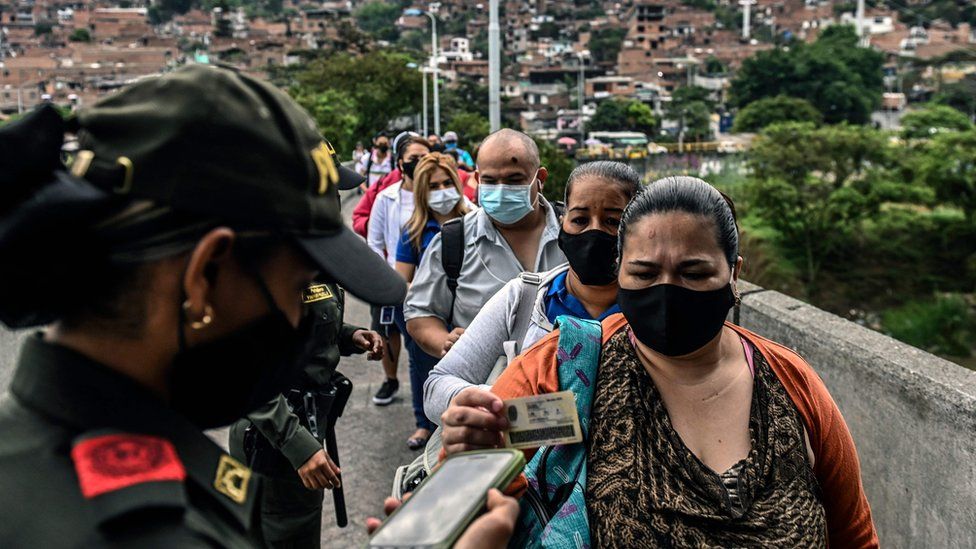 A queue of people wearing face masks in Medellin, Colombia. The woman at the front shows her ID to a police officer.
