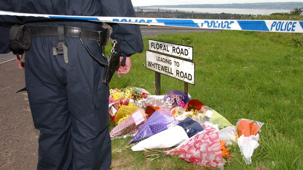 police officer next to floral tributes at murder scene