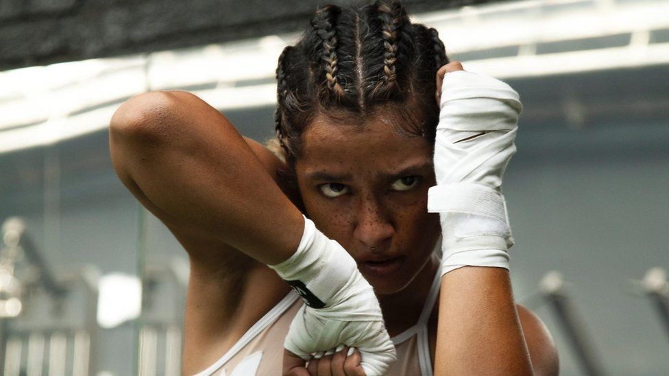 Ayesha Hussein. Ayesha is a 34-year-old British Asian woman. Her hair is braided and she poses in a fighting position, her left fist raised to her face while her right fist is pointed down. She has a freckled nose and brown eyes and an intense expression. She wears white gloves and a white gym top and she's pictured inside a gym.