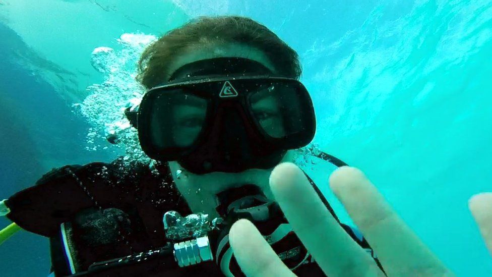 A woman underwater in diving goggles waves at the camera