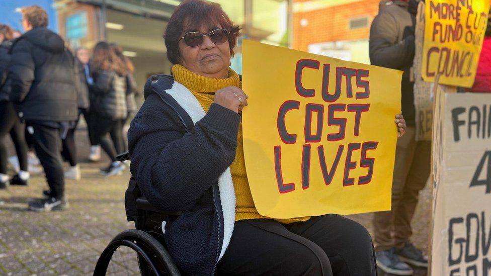 a woman in a wheelchair holding a placard saying cuts cost lives