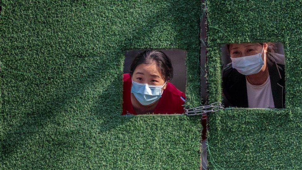 Женщины на карантине смотрят через маленькие окна в зеленом заборе на фоне блокировки Covid-19 в Шанхае, 2 мая 2022 г.