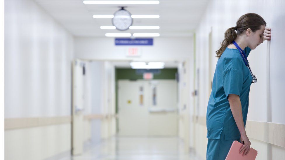 A stock image of a female doctor looking stressed. She is standing in a brightly lit hospital corridor, but has her left hand up against a wall and her head close up to the wall. She has her eyes closed.