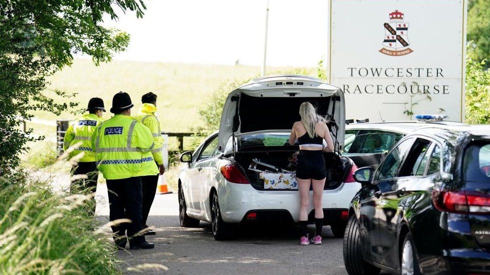 Three police officers watch a woman remove objects from her car boot
