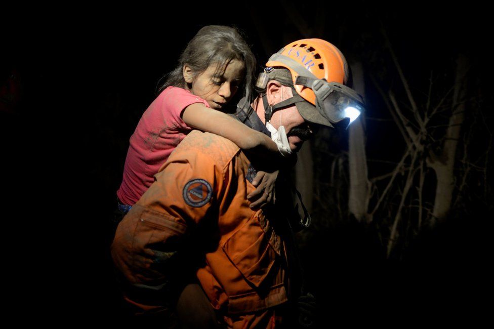 A rescue worker carries a child covered with ash