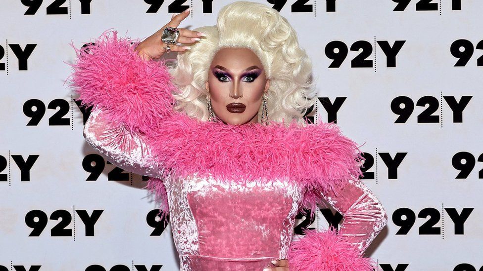 A drag queen in a pink feathered gown poses with a blonde wig
