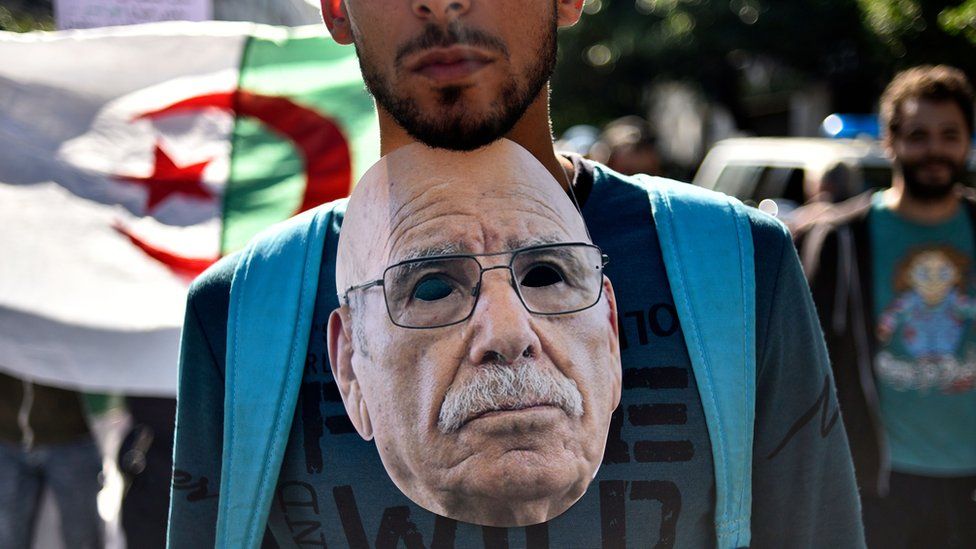 An Algerian protester hangs around his neck a mask representing Lakhdar Bouregaa, a well-known war veteran who was arrested by the authorities last summer, during an anti-government demonstration in the capital Algiers on November 1, 2019. - Demonstrators converged on Algiers in their thousands for a massive anti-government rally called to coincide with official celebrations of the anniversary of the war that won Algeria's independence from France.